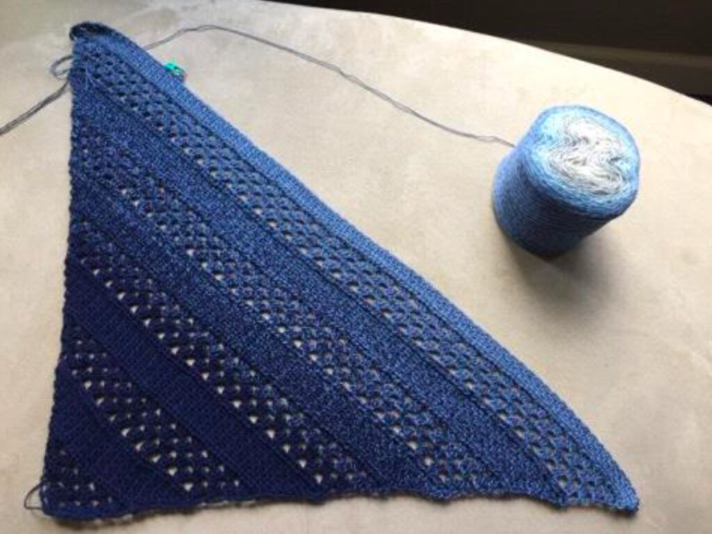 A blue and black crochet shawl laid out with a cake of yarn next to it.