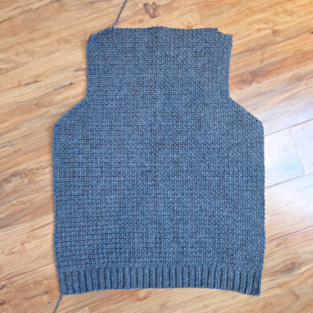 Working a blue grey colored crochet mens cardigan against a wooden background.