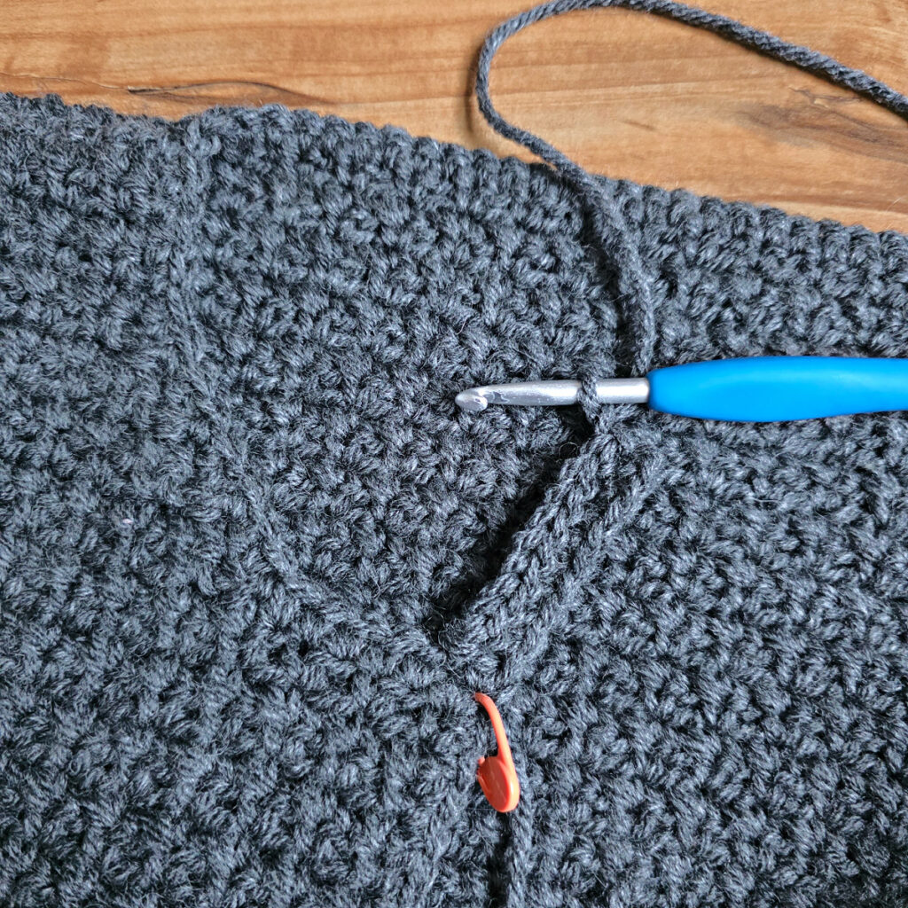 Working a blue grey colored crochet mens cardigan against a wooden background.