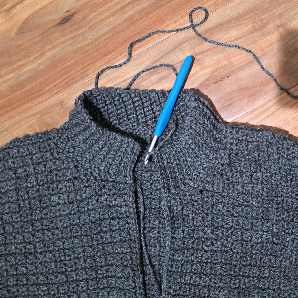 Working a blue grey colored crochet mens cardigan against a wooden background.