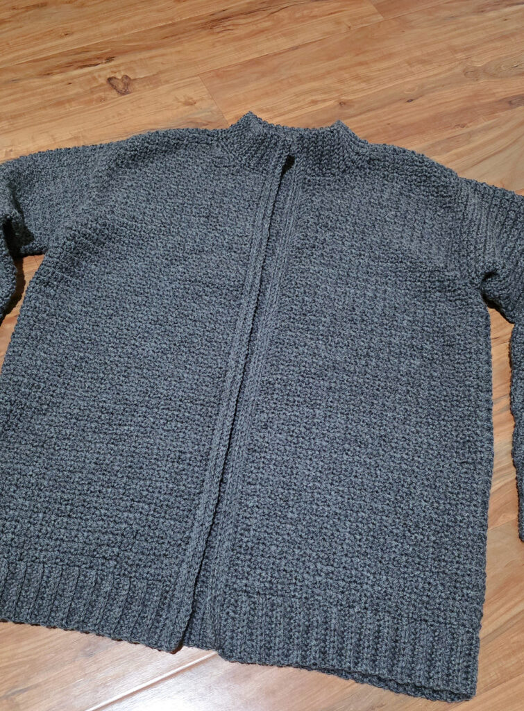 A blue grey colored crochet mens cardigan against a wooden background.