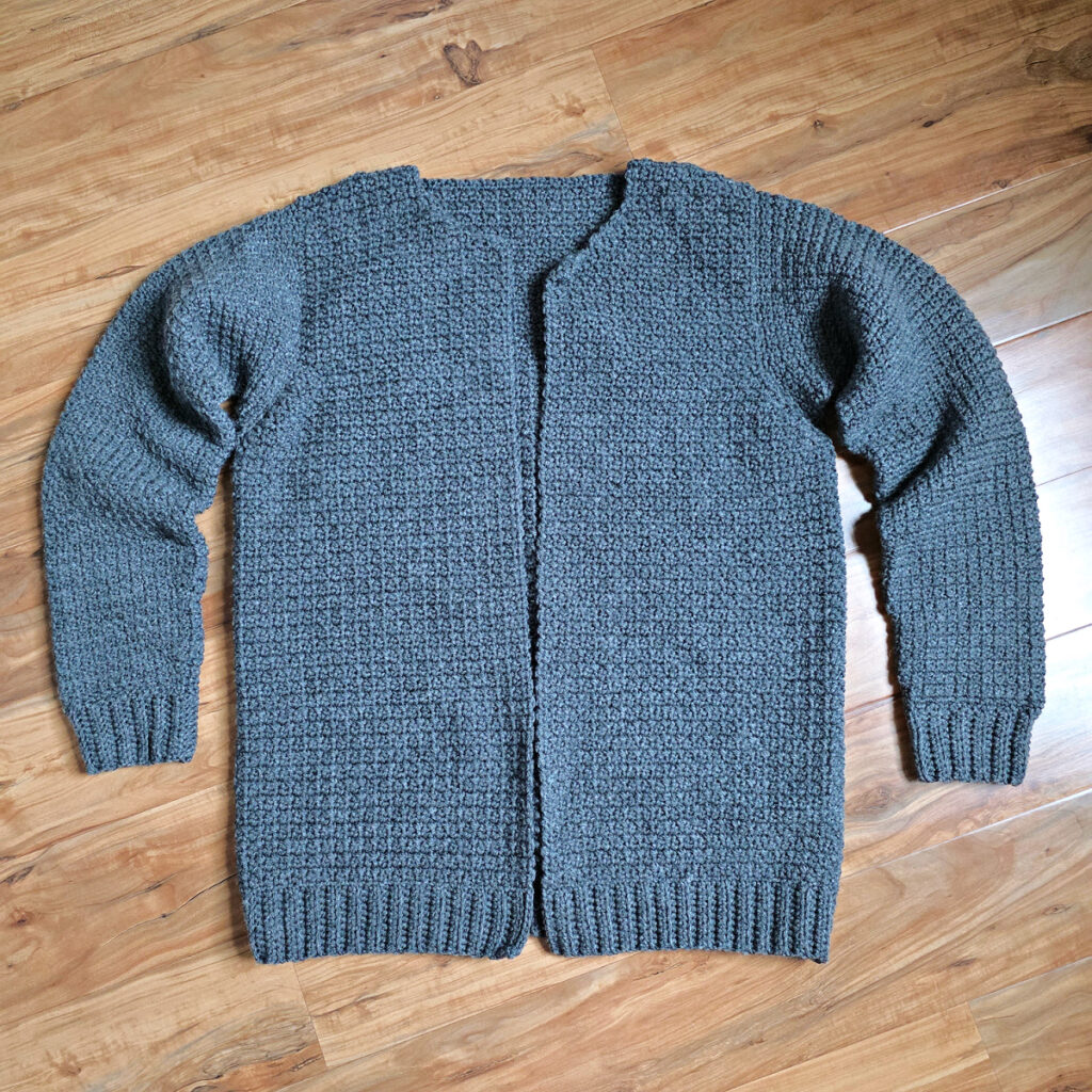 Working a blue grey colored crochet mens cardigan against a wooden background.