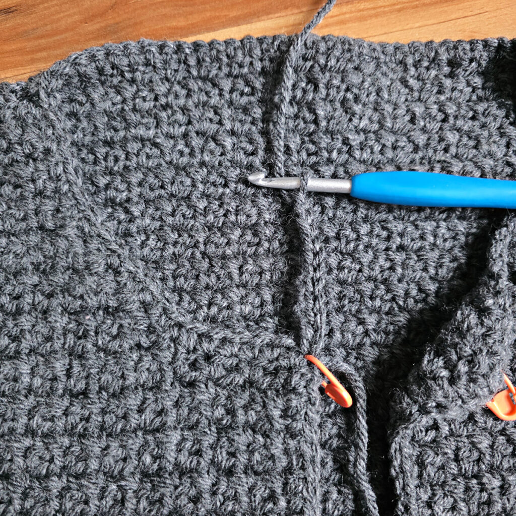Working a blue grey colored crochet mens cardigan against a wooden background.